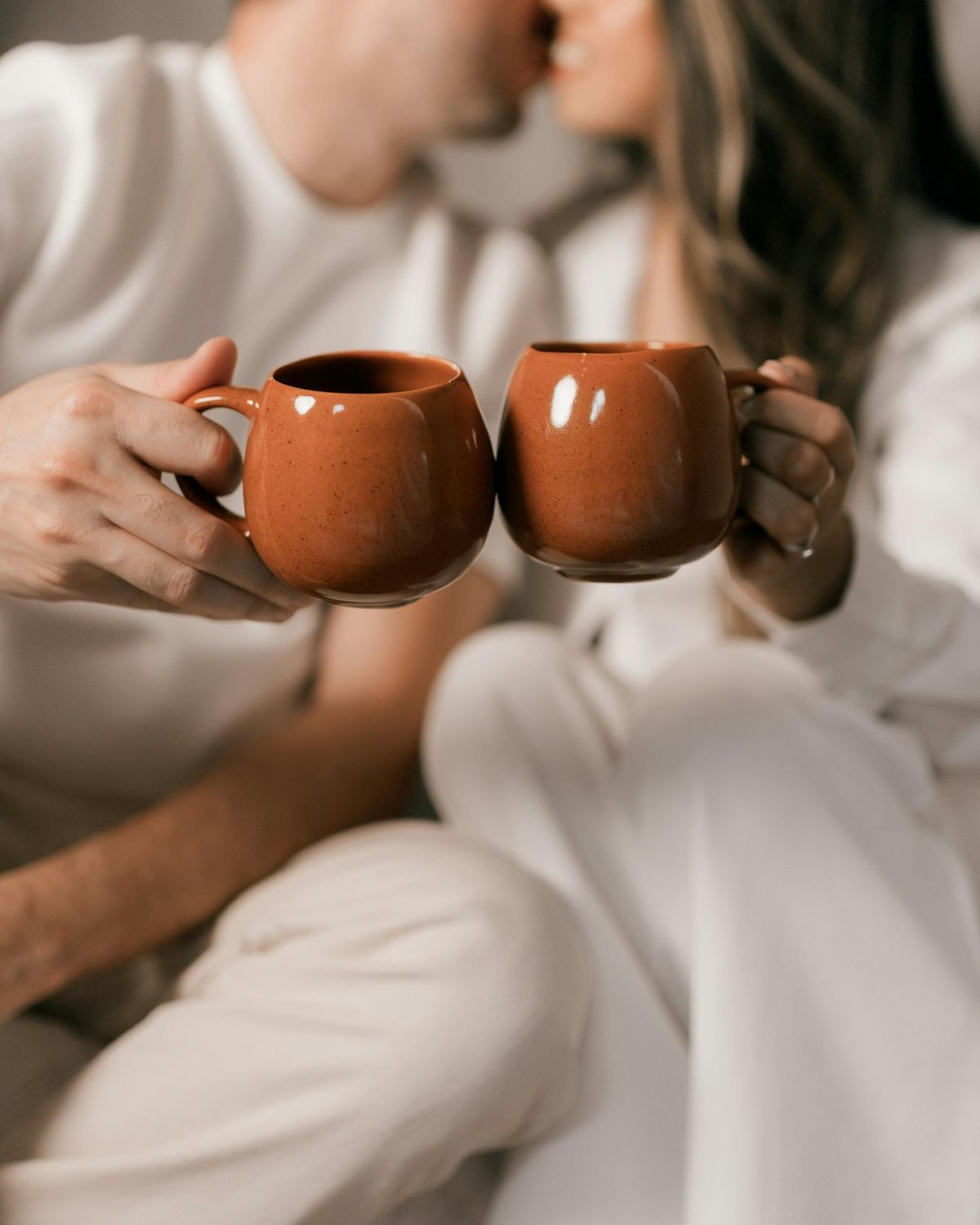 Two people in white sweaters clinking matching clay-colored mugs